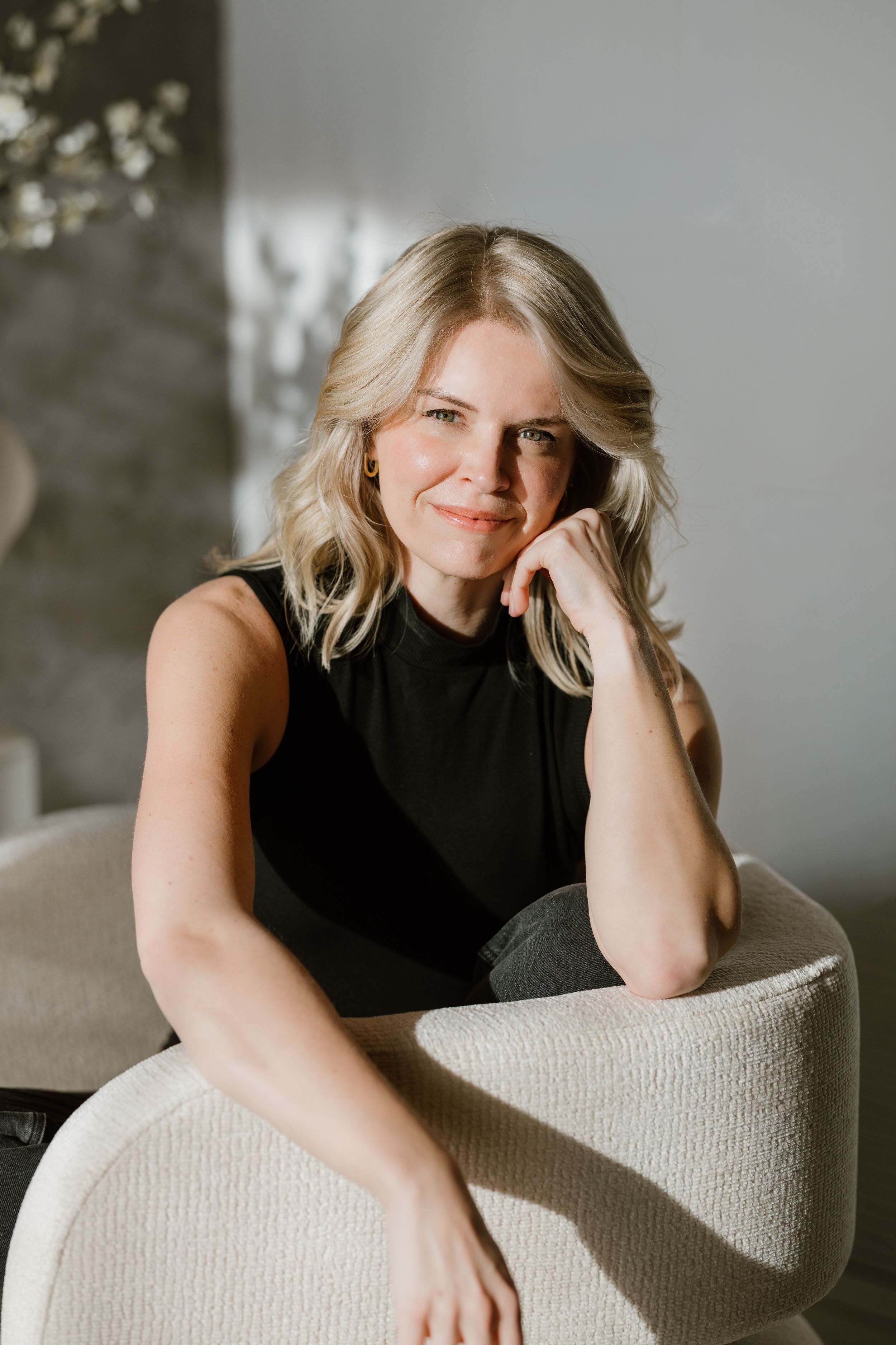 Smiling blonde woman in black sleeveless top sitting relaxed in a white chair in a modern interior.