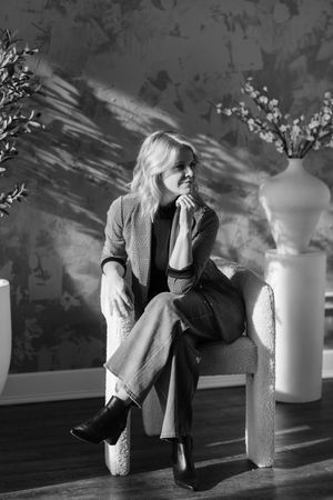 Black and white photo of a blonde woman sitting in a chair, wearing a blazer and wide-leg pants with ankle boots.