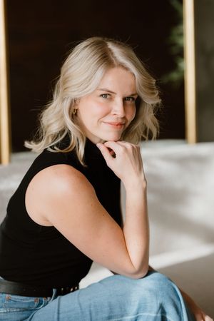 Blonde woman in black sleeveless top and jeans sits posed with hand near chin, smiling softly indoors.