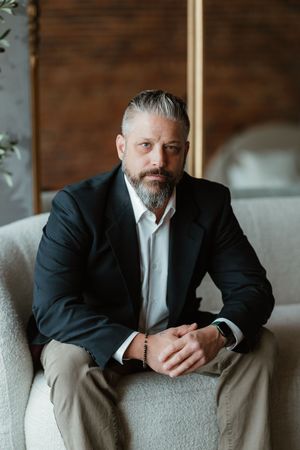 Professional portrait of person in dark suit with gray beard sitting casually against rustic background.