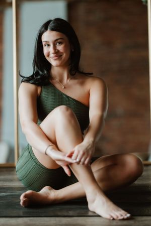 Relaxed woman in olive green one-shoulder outfit sits on wooden floor with natural lighting and rustic background.