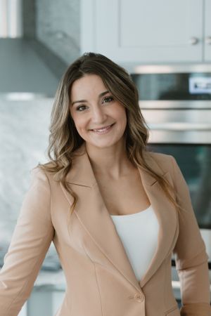 Professional portrait of person in tan blazer and white top in modern kitchen.
