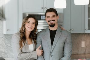 Professional portrait of couple in business attire standing in modern white kitchen.