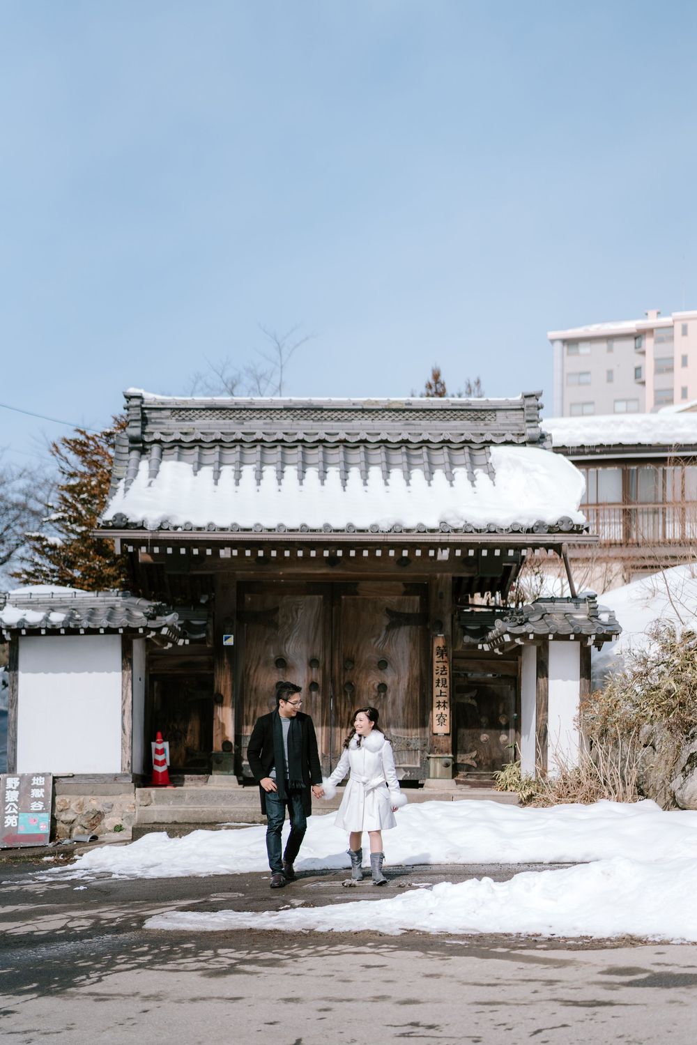 Couple holding hands walking in front of a snow-covered traditional Japanese gate in winter.
