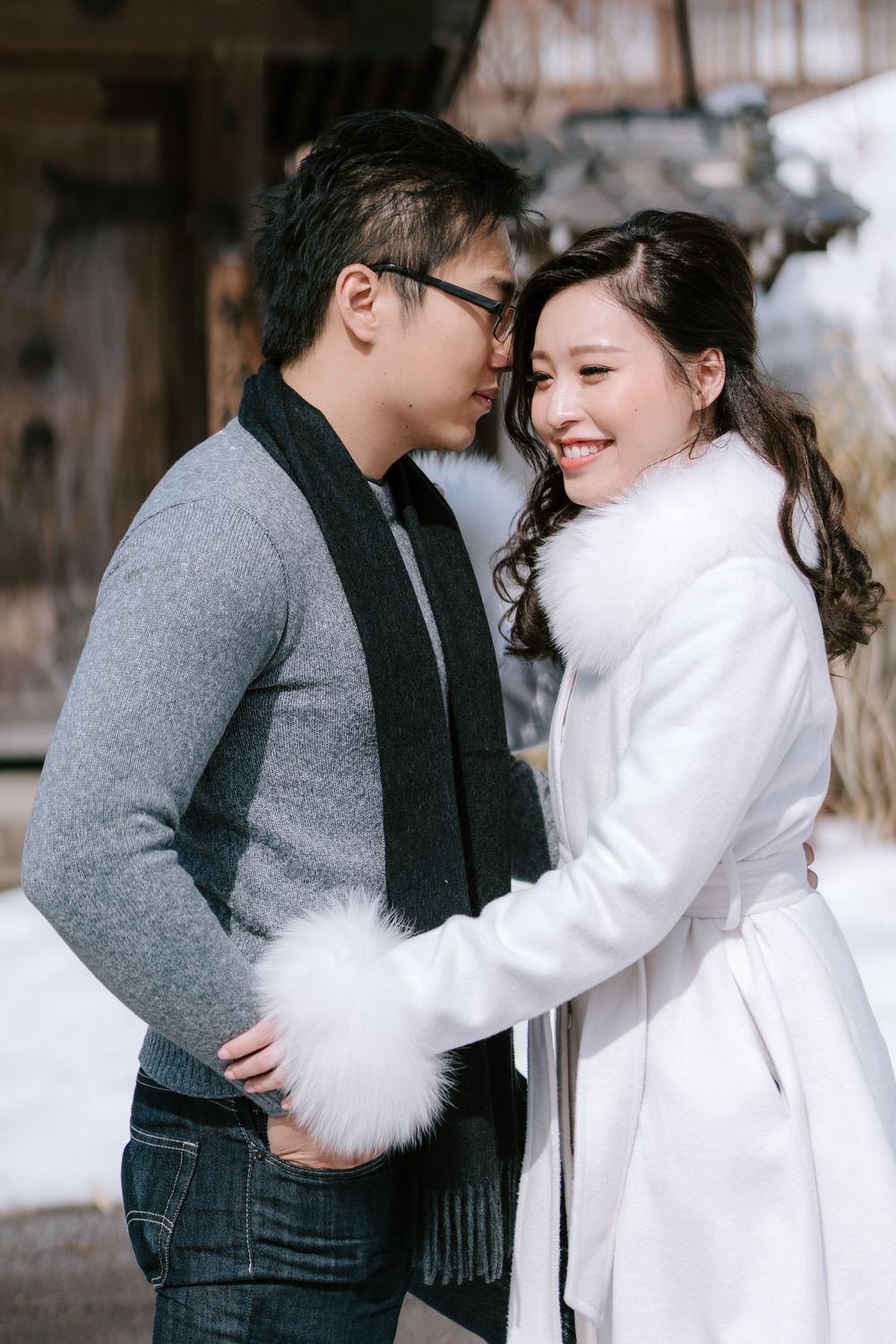 A smiling couple in winter attire embraces outdoors in the snow, woman in white fur coat, man in gray sweater.