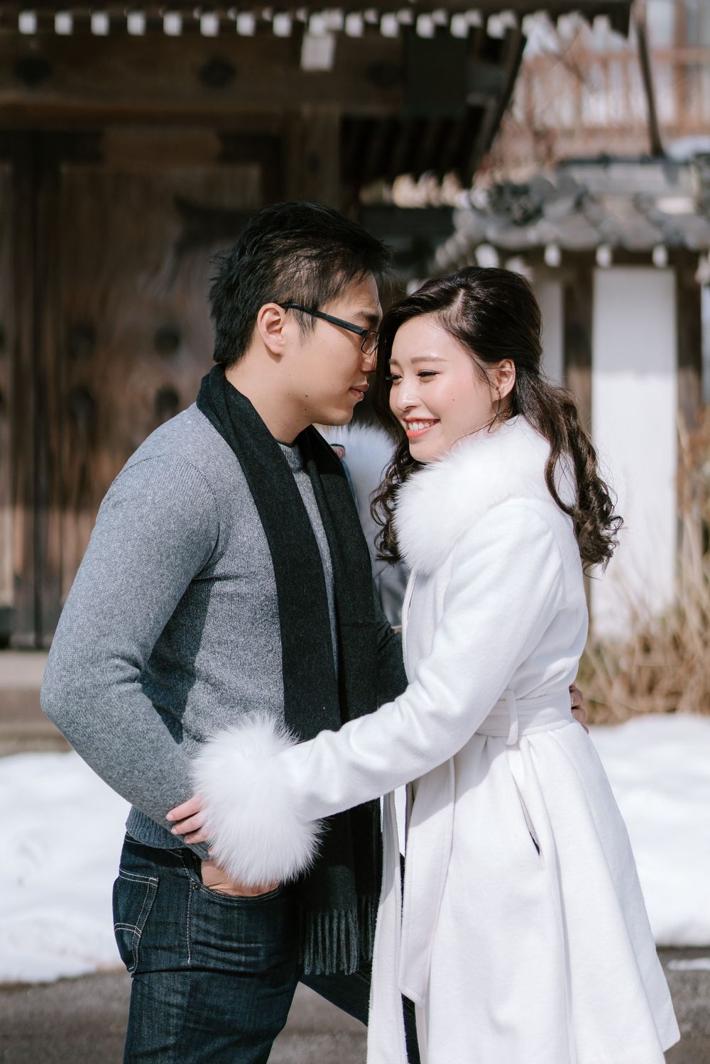 Couple embracing in snow wearing winter outfits, woman in white coat smiling, man in gray sweater and scarf, traditional building backdrop.