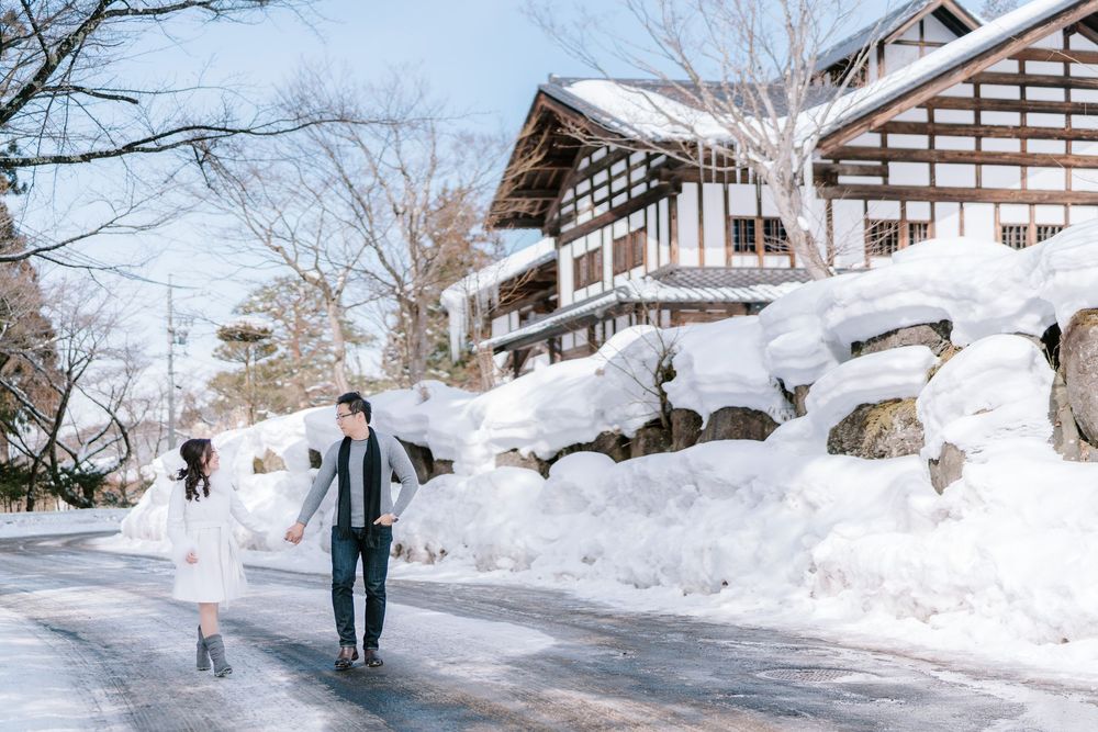 Couple holding hands on a snowy road beside a traditional Japanese building with large snow drifts in winter.