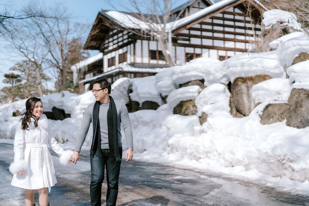 Couple walking hand in hand in winter snow landscape near traditional Japanese building on sunny day.