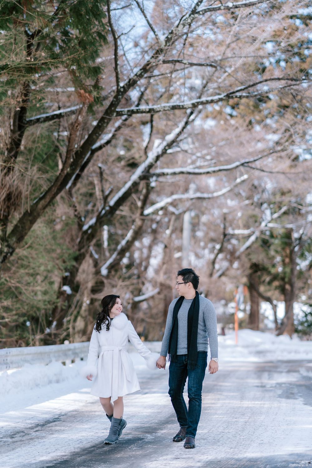 Couple holding hands walking on snowy road surrounded by winter trees.