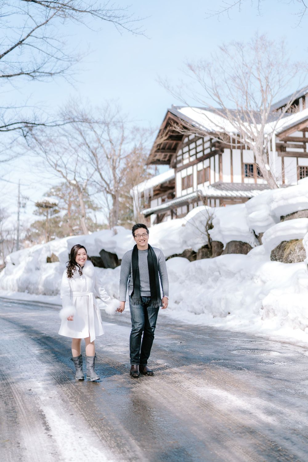 Couple walking hand in hand on a snowy road in Japan, with traditional architecture and deep snow banks behind them.