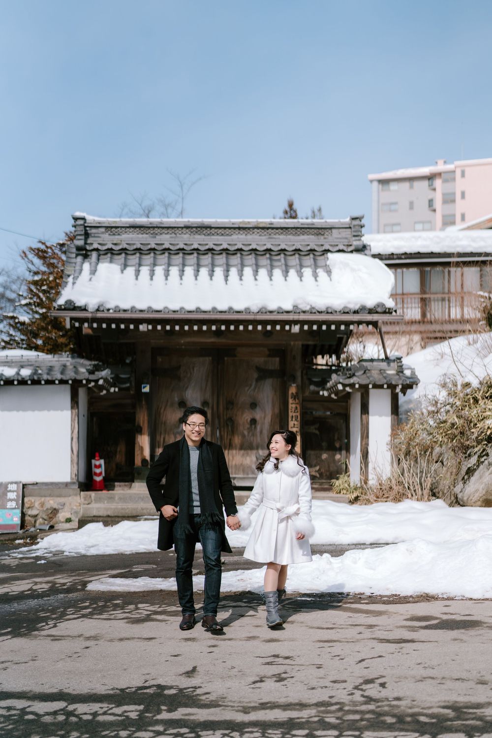 Happy couple strolling hand in hand before a snow-laden Japanese architectural gate on a sunny day.