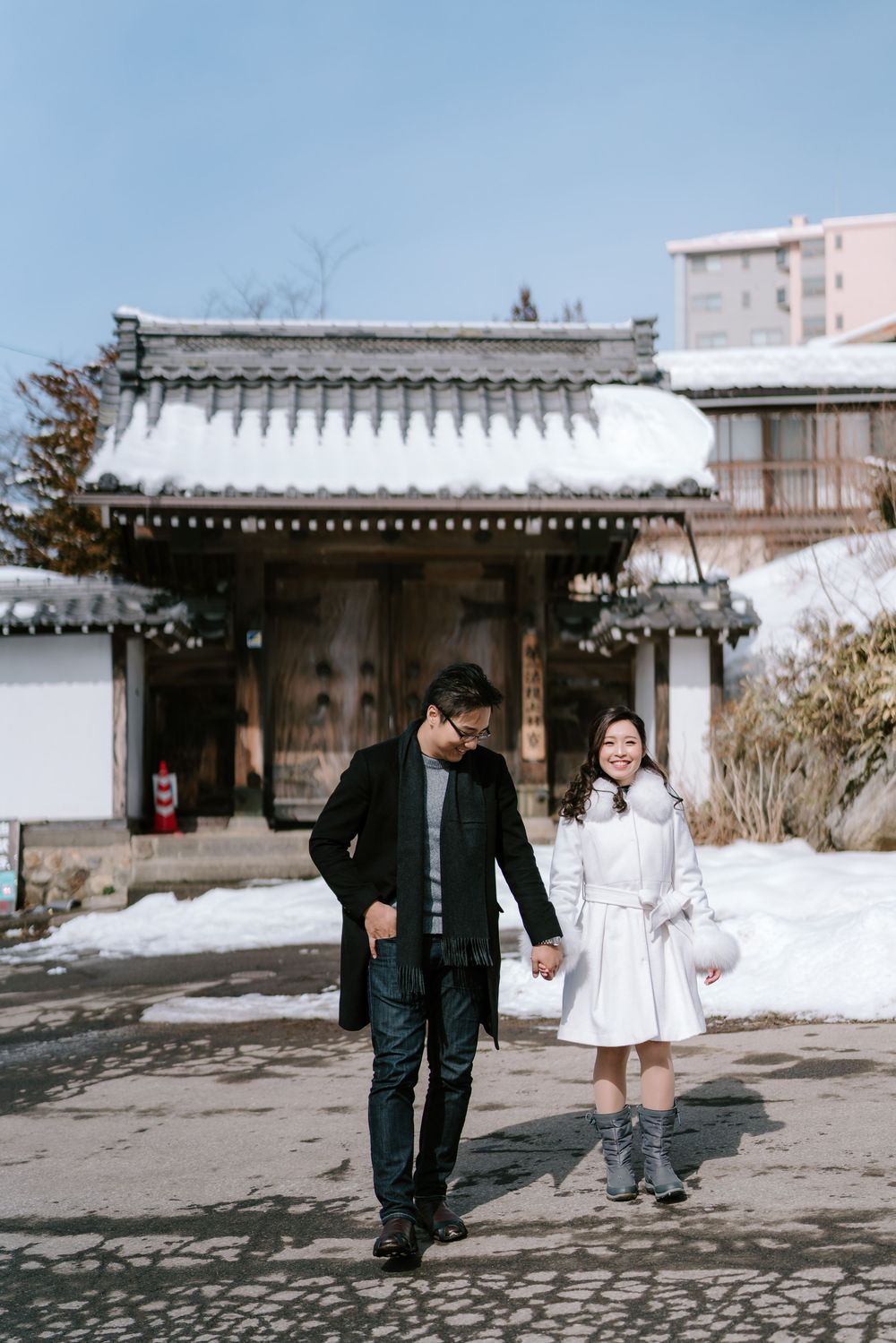 Couple holding hands walking in front of a snow-covered traditional Japanese gate on a sunny winter day.