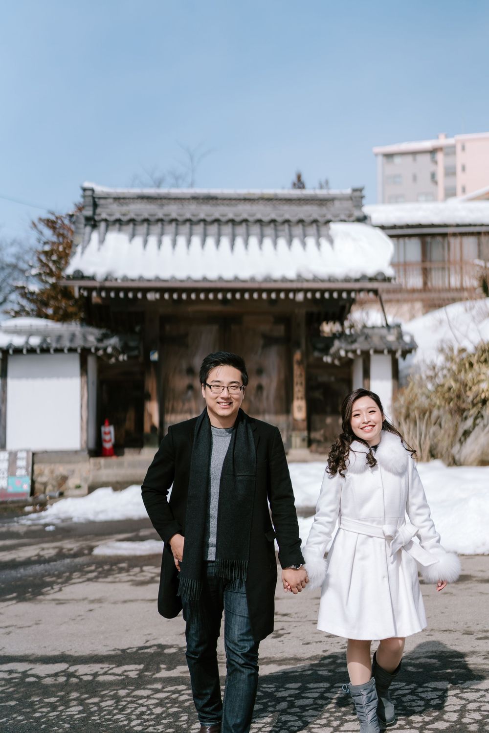 Couple holding hands walking in front of a snow-covered traditional Japanese building on a sunny winter day.