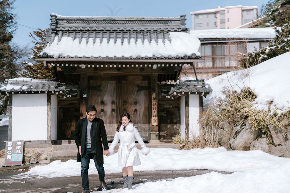 Couple holding hands walking in snow before a traditional Japanese gate with heavy snow on the roof.