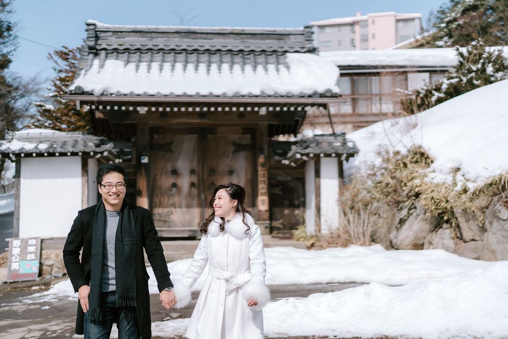 Couple holding hands in snow outside traditional Japanese gate, woman in white dress, man in black coat, winter wedding photo.