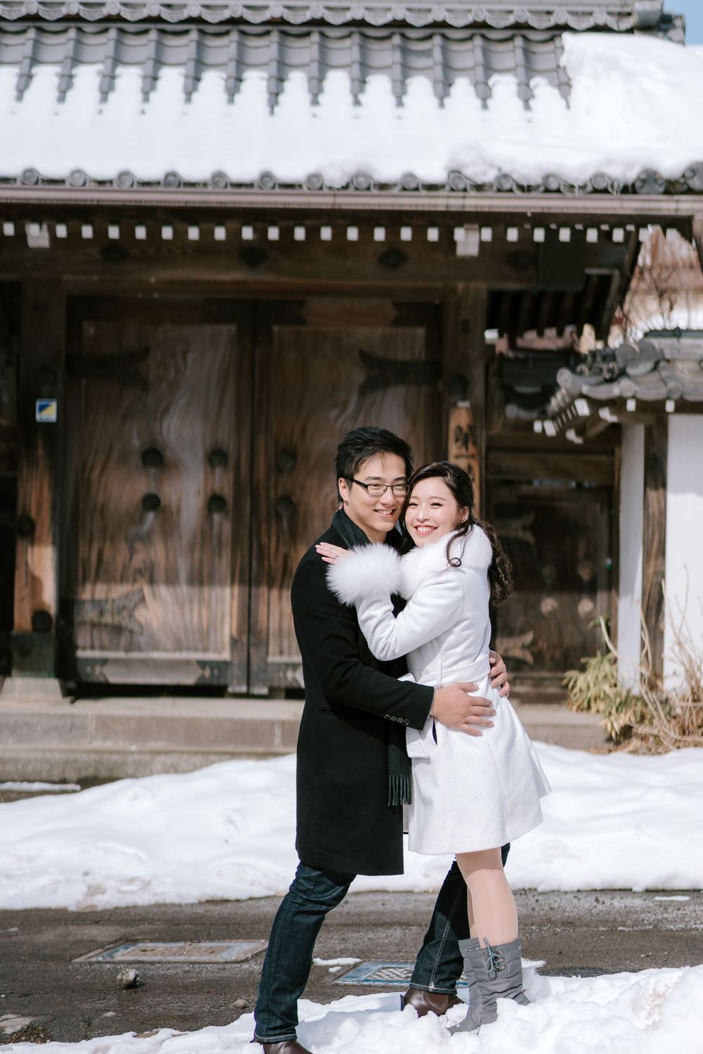 Couple embracing and smiling in the snow outside a traditional Japanese building in winter.