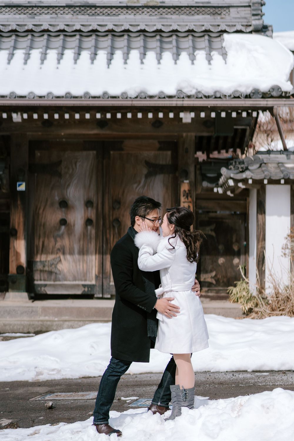 Couple sharing a romantic kiss in the snow outside a traditional Japanese building in winter.
