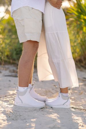 Close-up view of feet wearing matching white shoes standing on sandy beach during a sunny day.