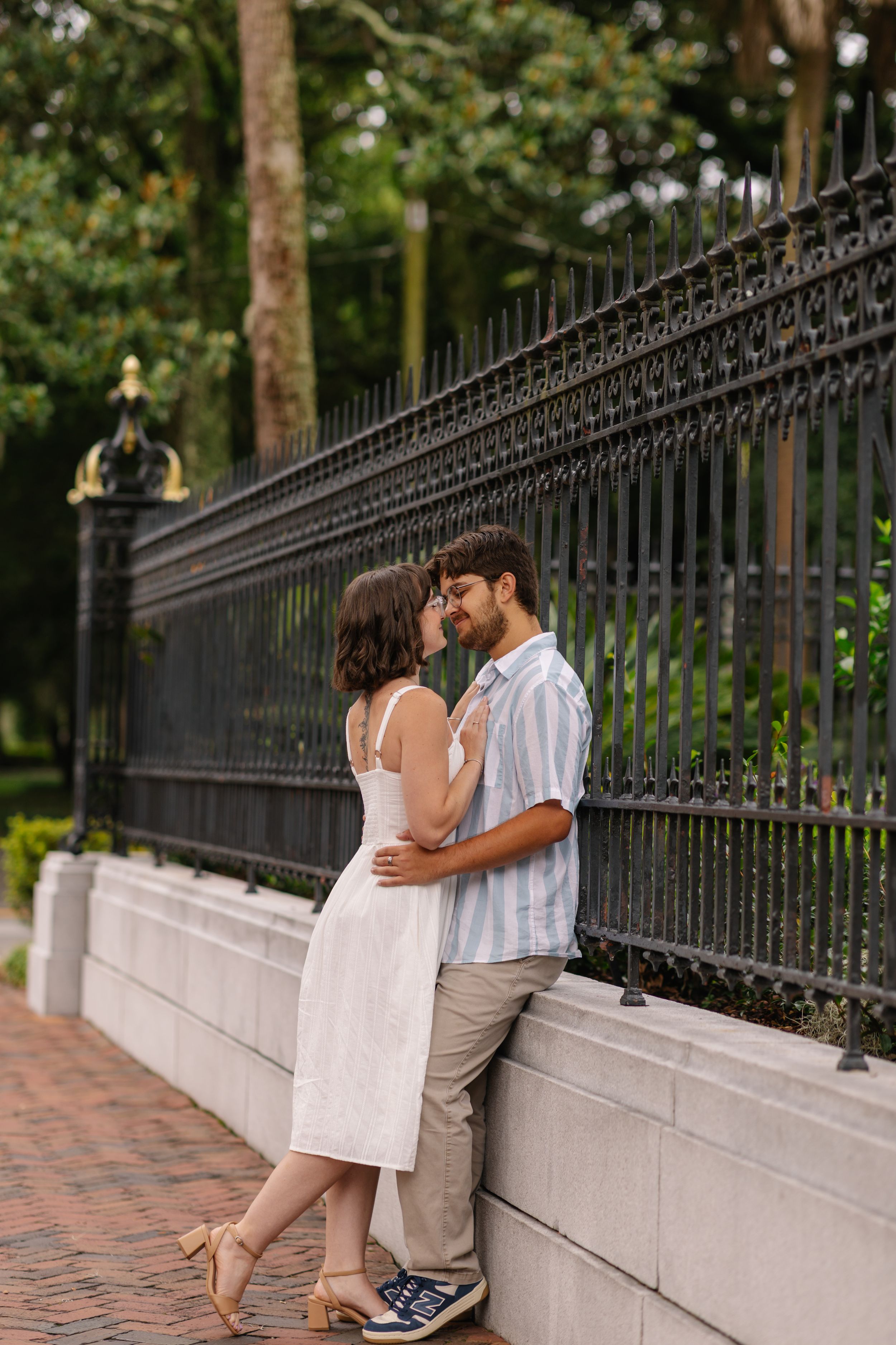 A couple shares an intimate moment while sitting on a concrete wall next to a decorative iron fence in a park setting.