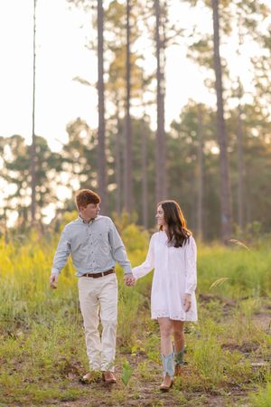 A couple walks together through a sunlit pine forest while wearing white and holding hands.