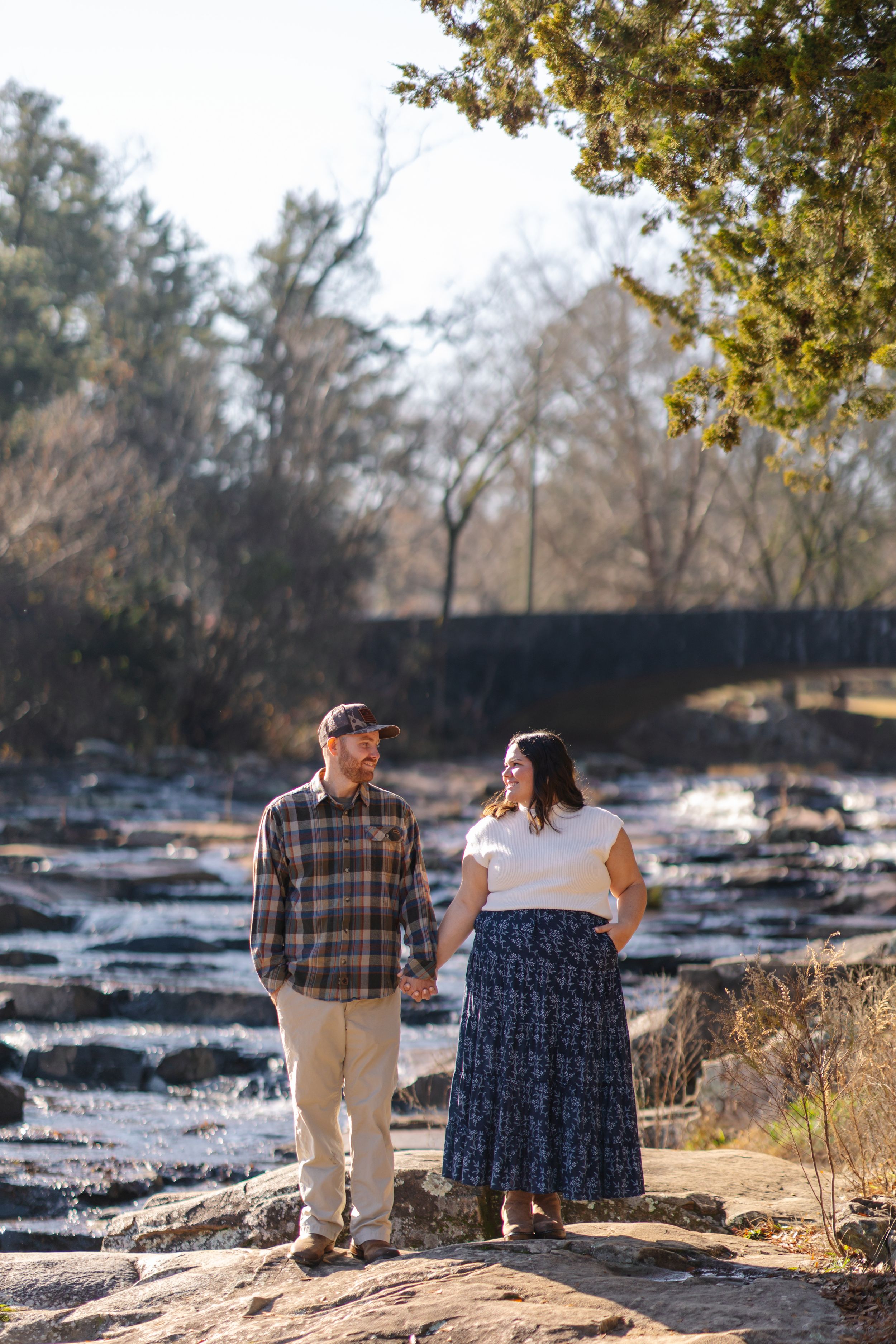 A couple sits together on rocks by a river during a sunny autumn day, enjoying the peaceful natural scenery.