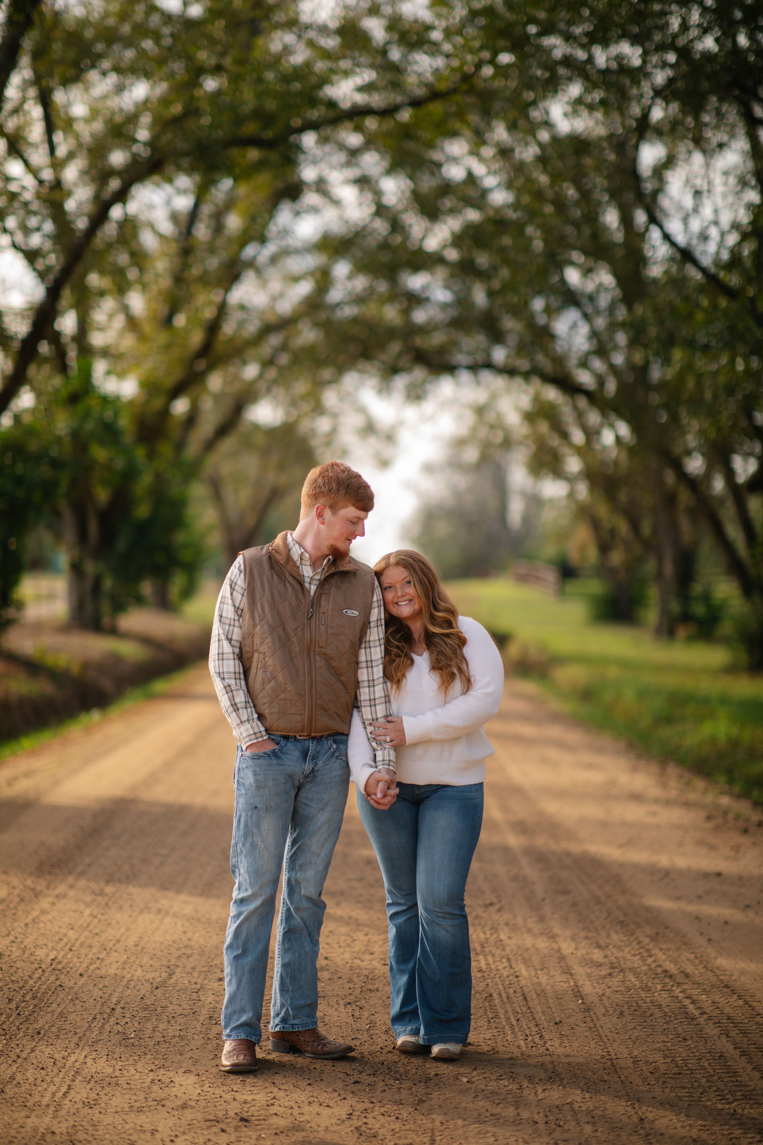 A couple walks together on a dirt path surrounded by trees in a romantic outdoor setting.