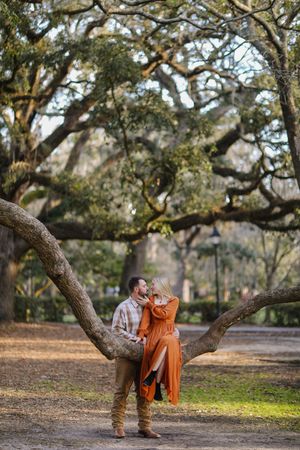 A person in an orange dress sits on a curved tree branch in a peaceful park setting during autumn.