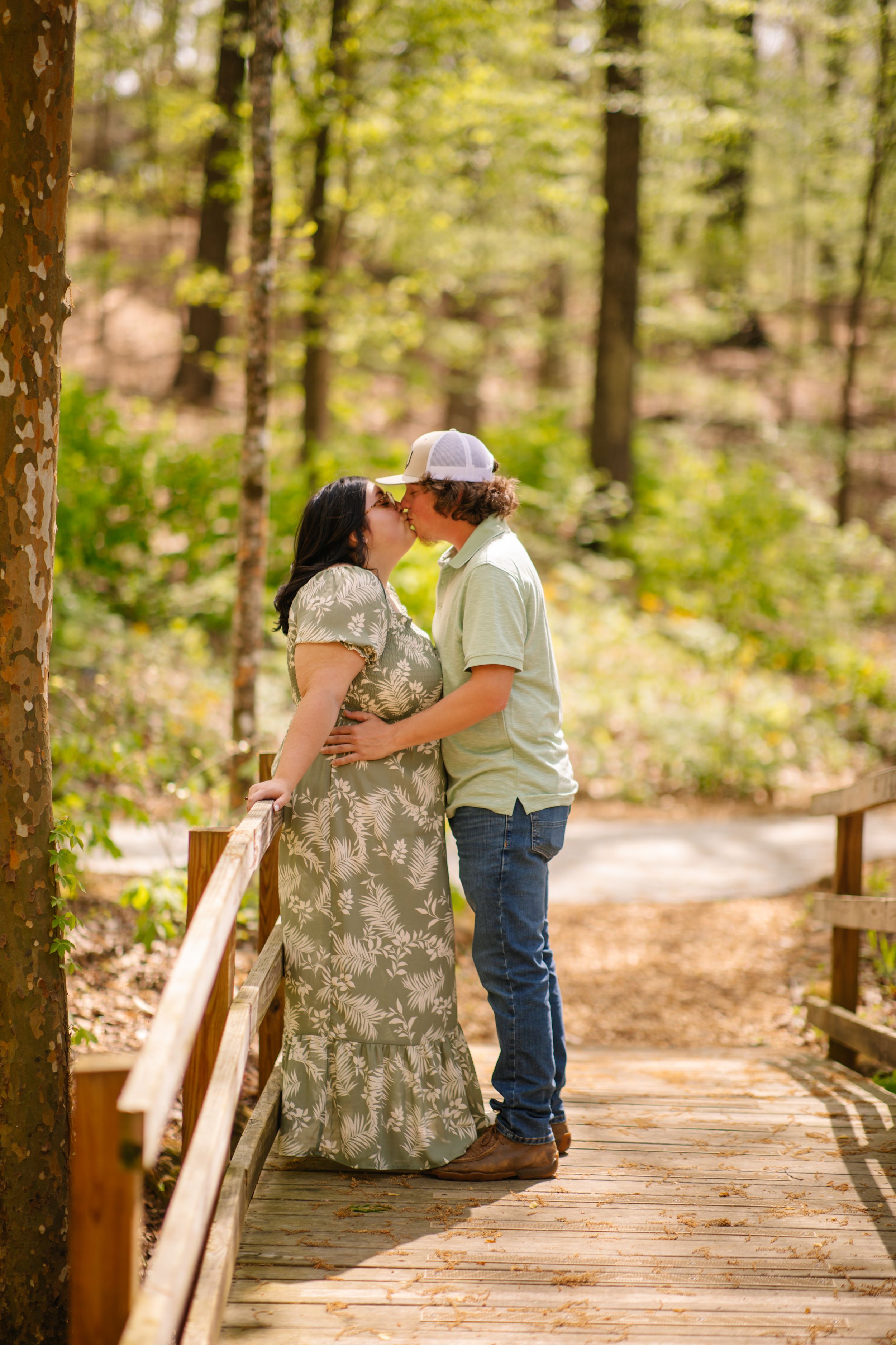 A couple shares a tender moment while sitting together on a wooden boardwalk trail through a sunlit forest.