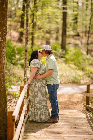 A couple shares a tender moment while sitting together on a wooden boardwalk trail through a sunlit forest.