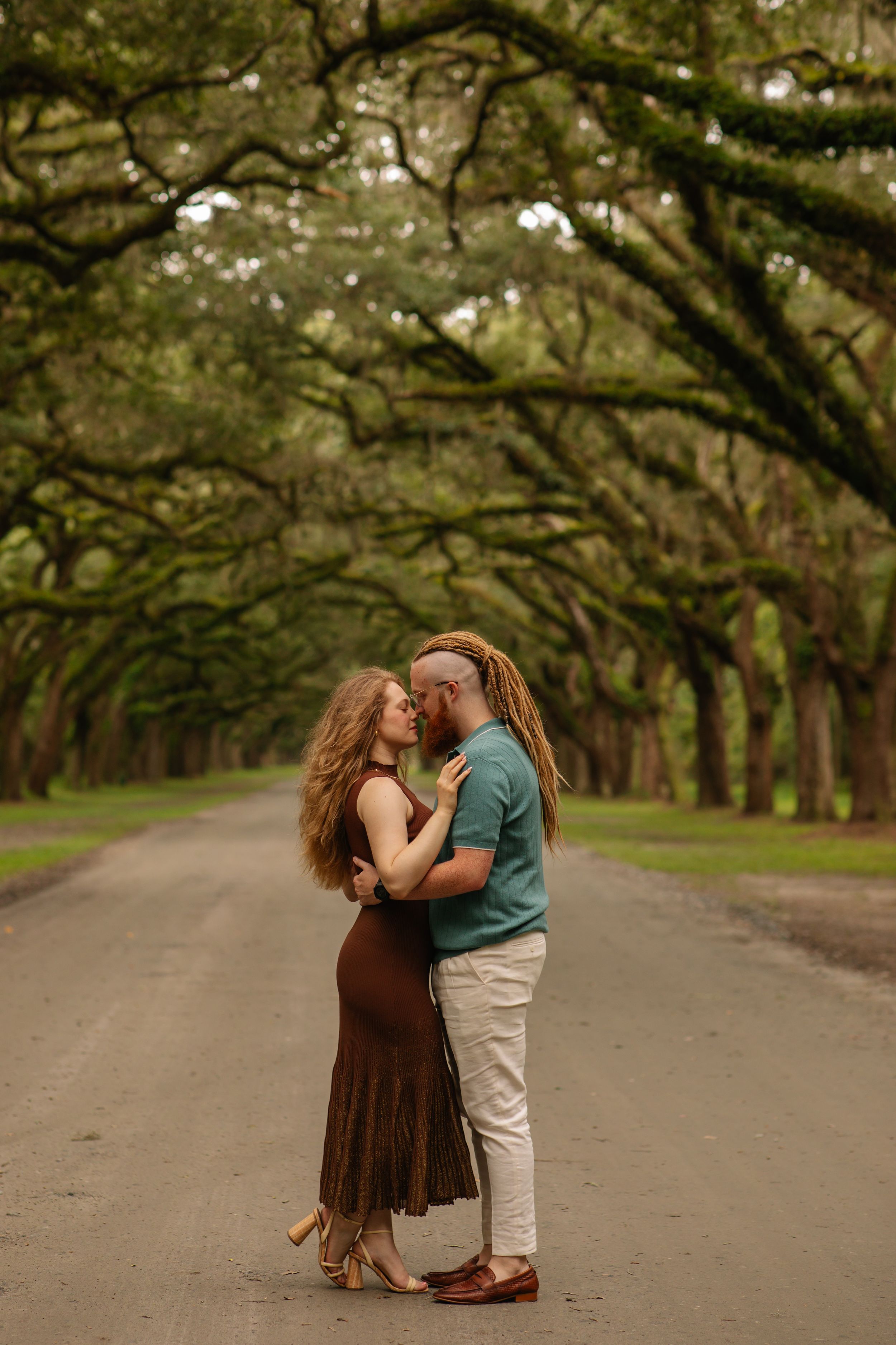 A couple shares romantic moments under a canopy of oak trees along a scenic path in a park.