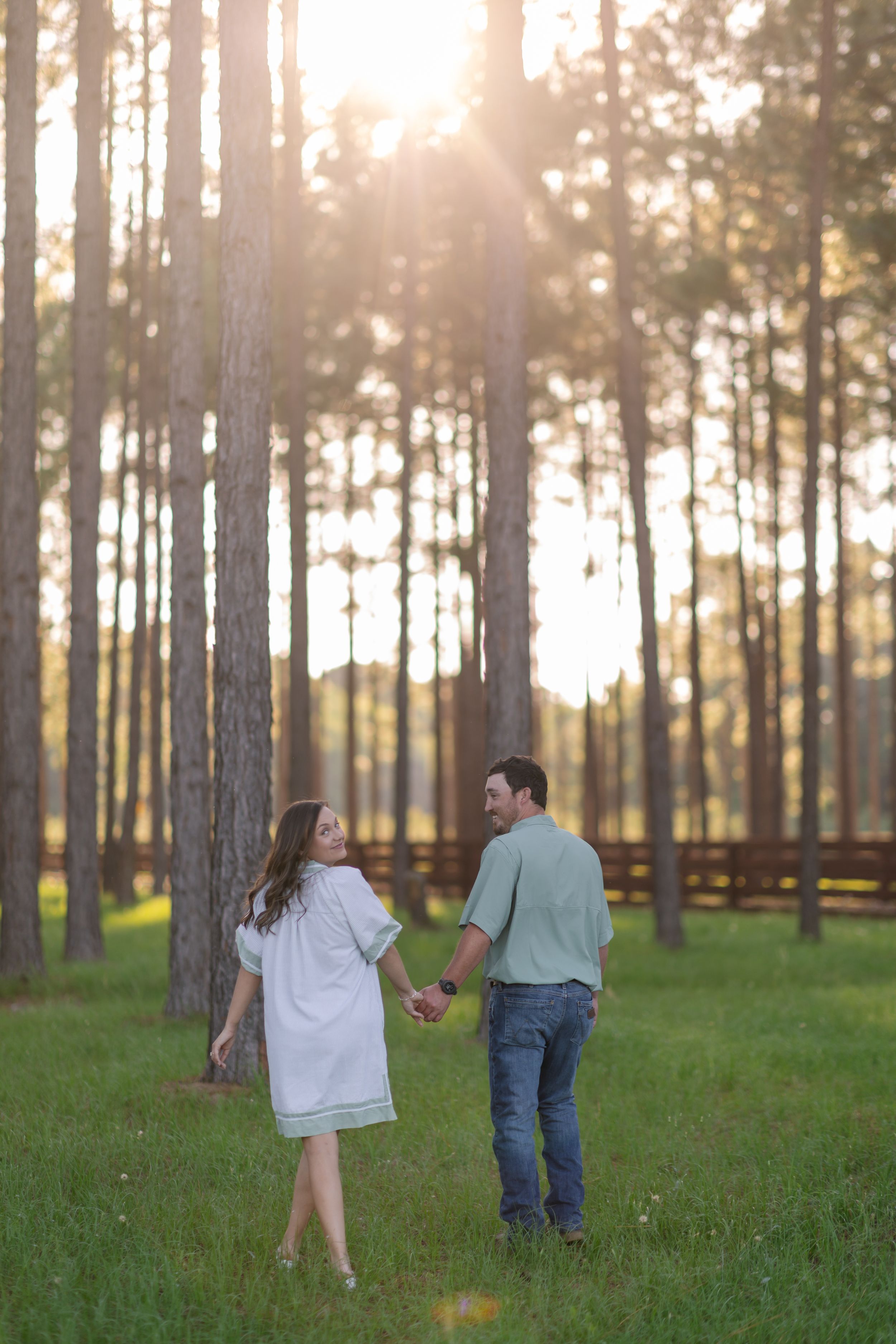 Couple holding hands in a sunlit pine forest, woman in white dress and man in green shirt and jeans.