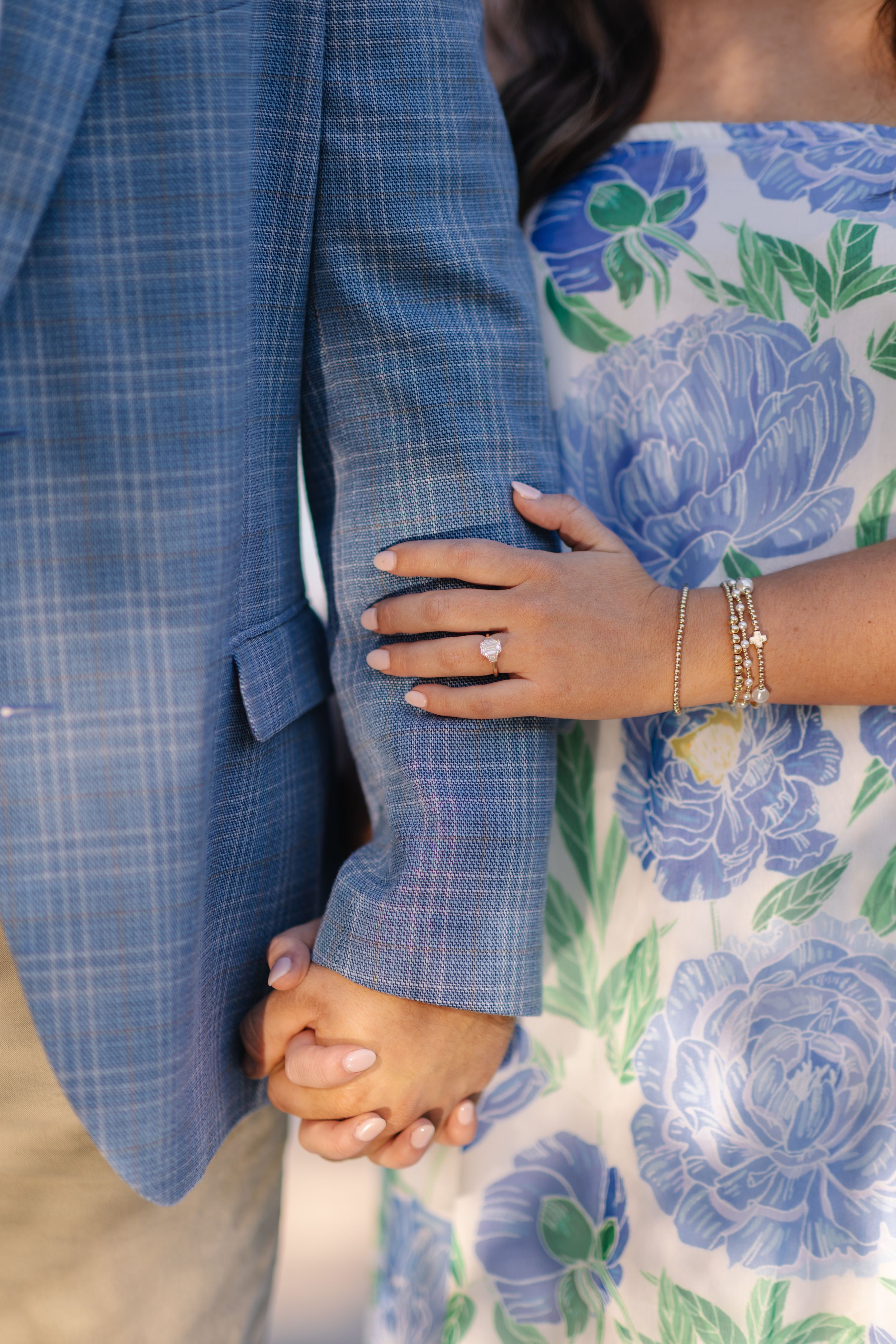 Couple holding hands, woman wearing diamond engagement ring and bracelet, blue floral dress beside man in plaid suit.