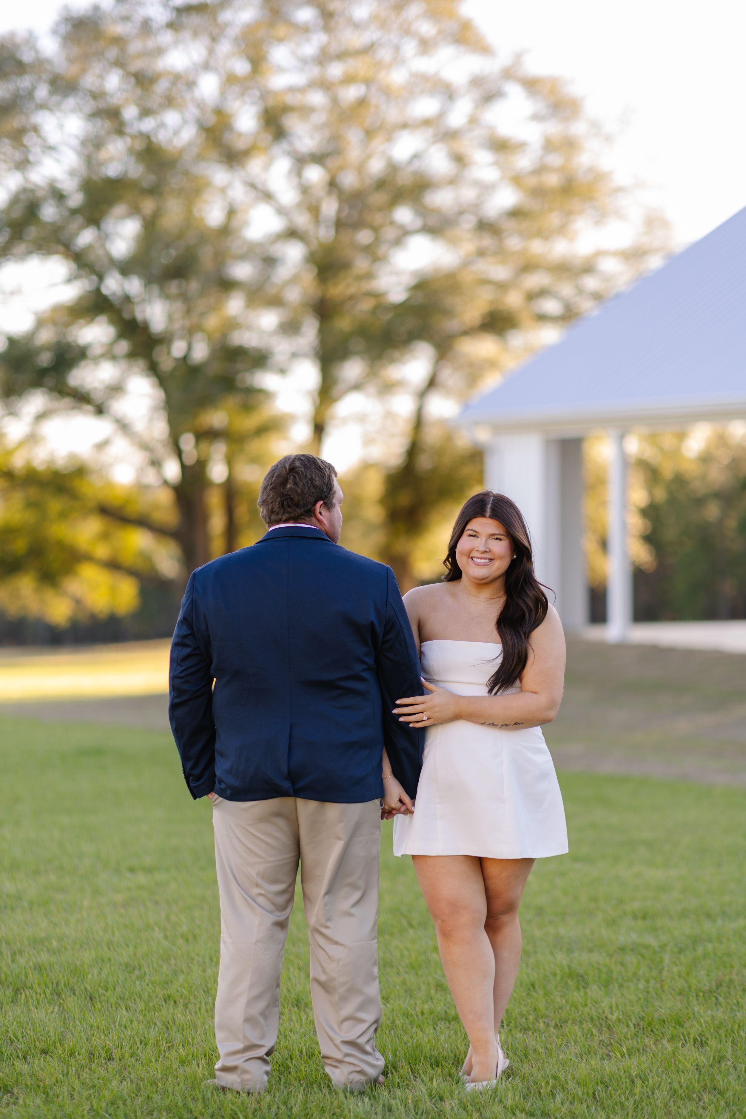 Couple holding hands on green lawn, woman in white mini dress smiling, man in navy blazer, golden hour light.