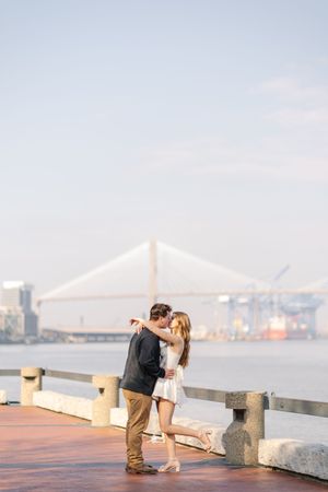 Couple sharing a romantic moment on a waterfront boardwalk with a suspension bridge visible in the background.