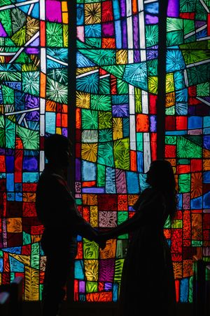 Silhouettes of people stand against a vibrant stained glass window backdrop with colorful geometric patterns and designs.