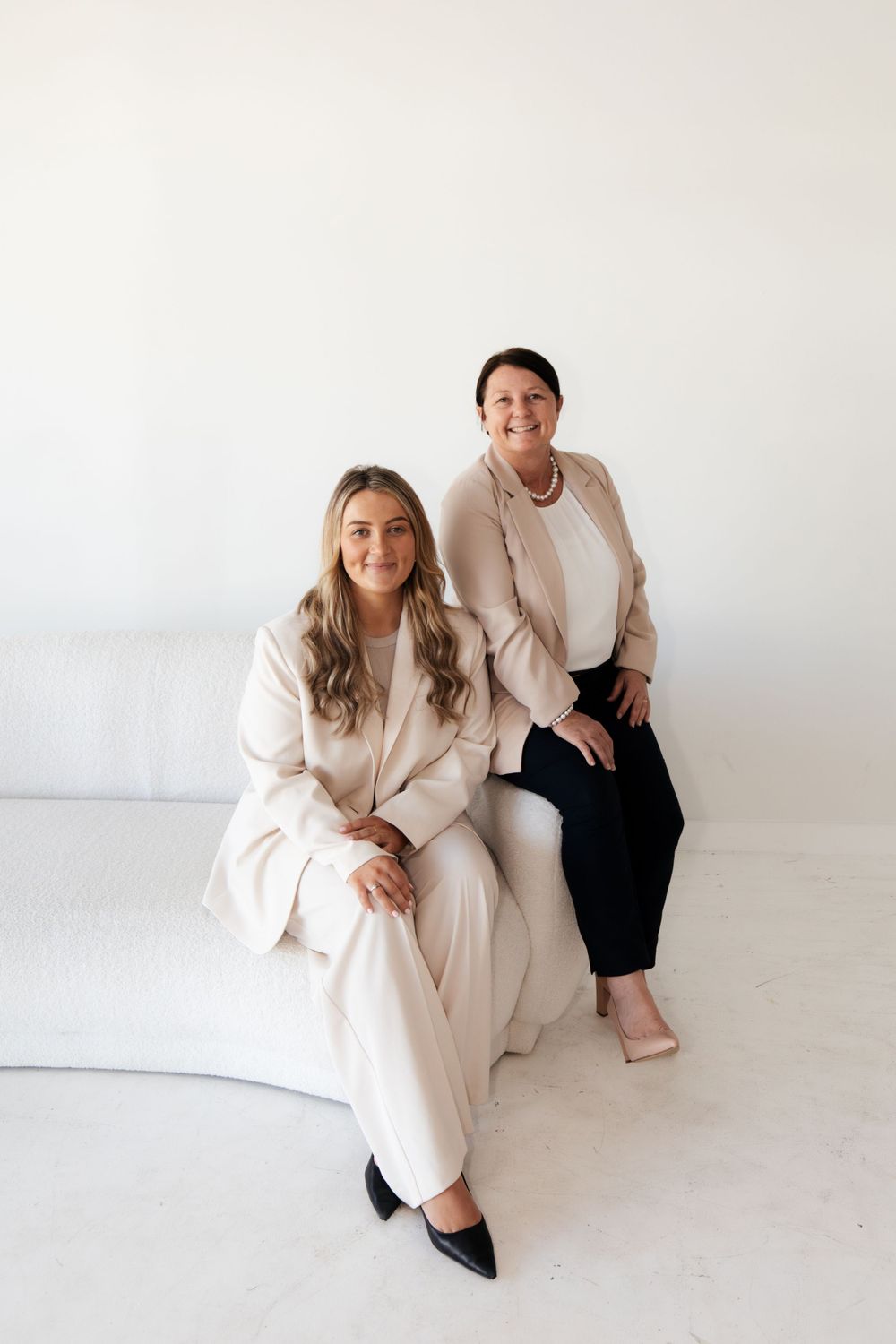 Two colleagues pose together on white couch in cream and black business attire.