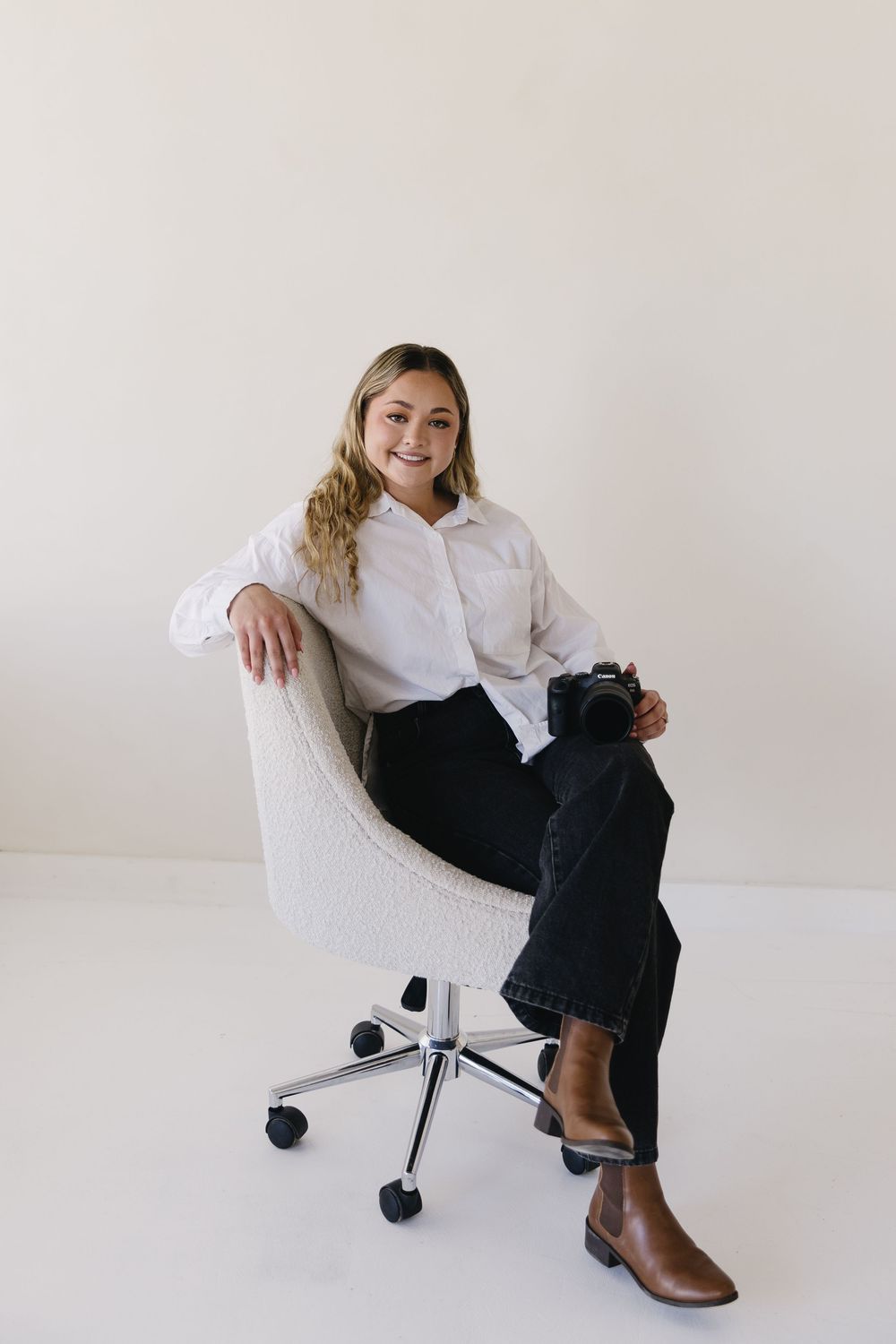 Professional seated on white office chair wearing classic white button-up shirt, dark pants and brown boots against neutral background.