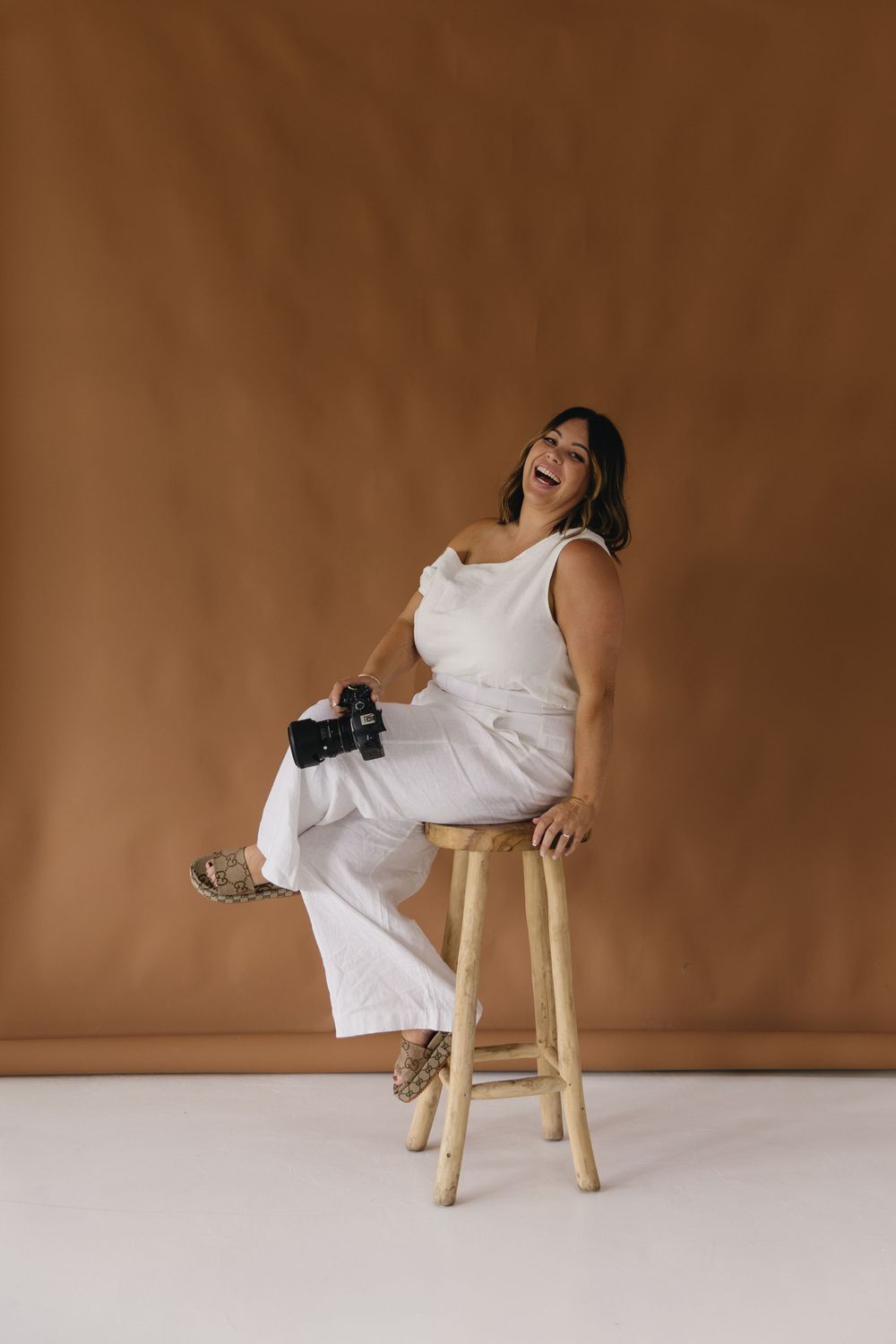 Joyful photographer in white outfit sits on wooden stool with camera against brown backdrop in studio setting.