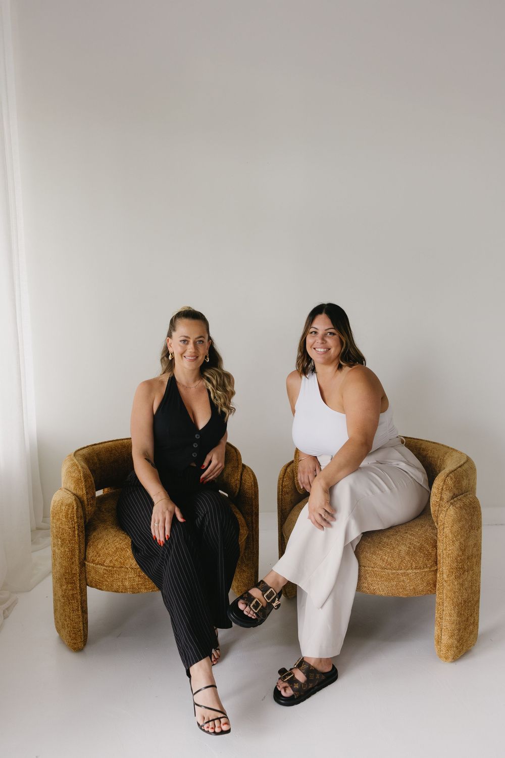 Two women sit together on a vintage gold velvet curved sofa wearing contrasting black and white outfits against white backdrop.