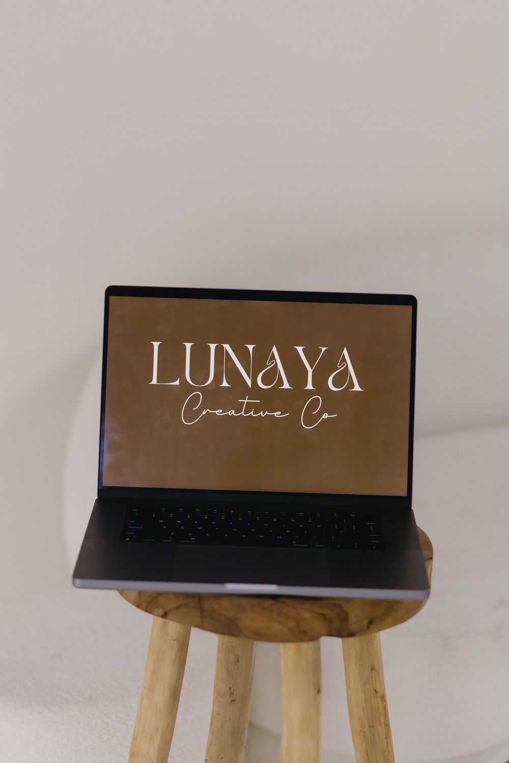 Laptop displaying Lunaya Creative Co. logo on screen sitting on wooden stool against white background.