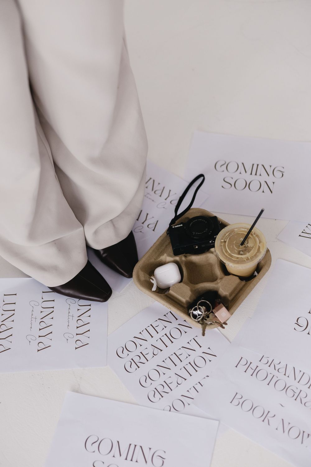 Minimalist composition with black shoes, coffee cup on gold tray, and 'Coming Soon' text papers on white background.