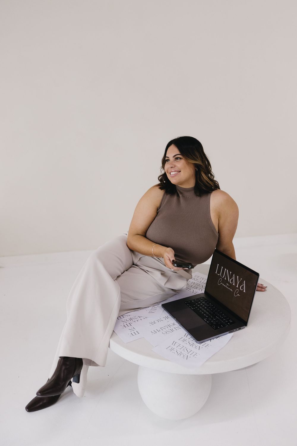 A person in brown sleeveless top working on laptop while lounging on white furniture in minimalist setting.