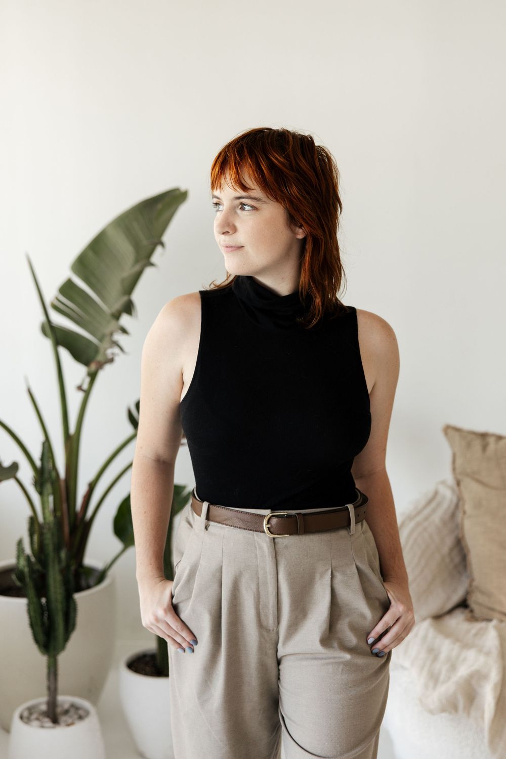 A stylish outfit featuring a black sleeveless turtleneck and tan high-waisted trousers with a houseplant in the background.