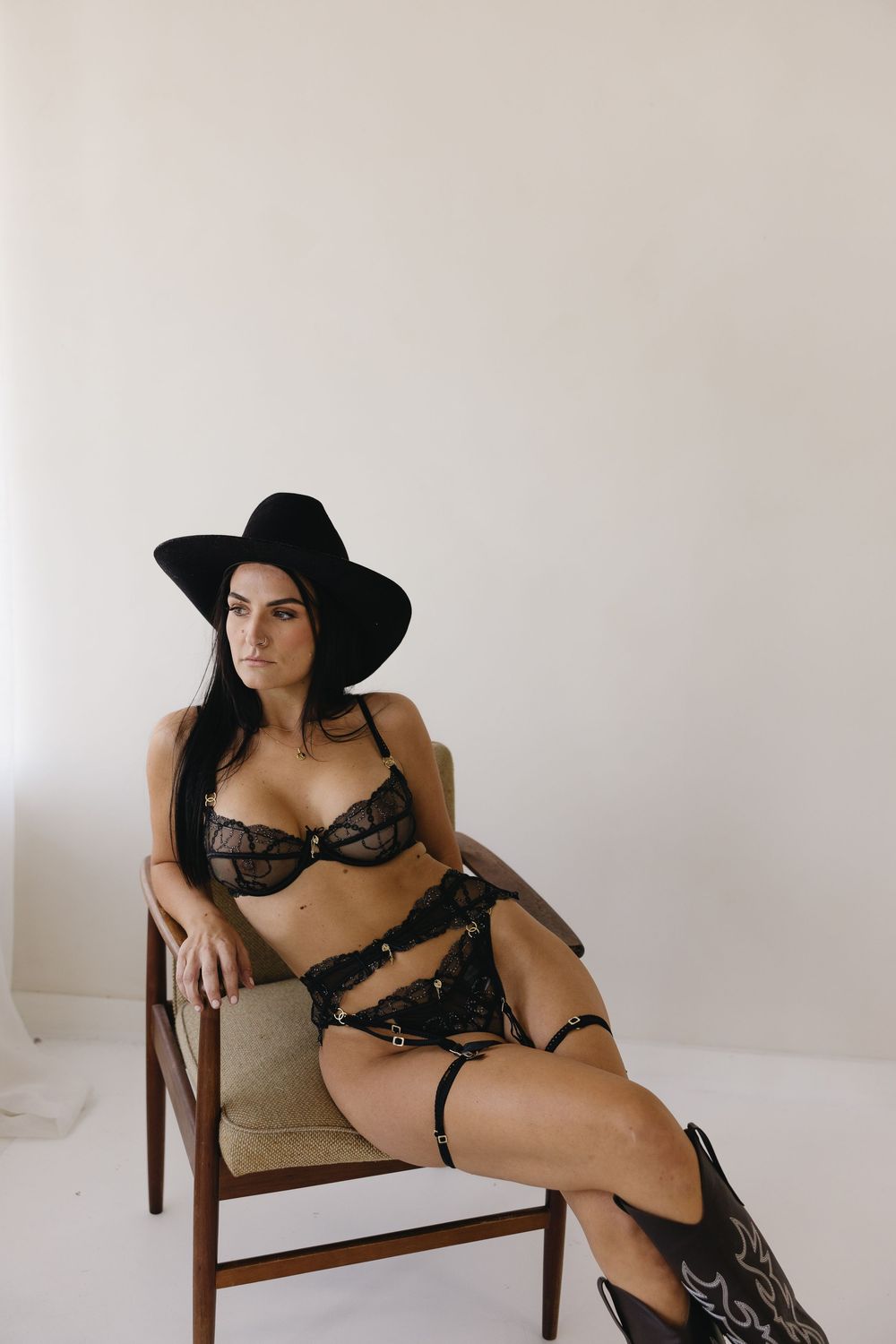 Figure in black hat and dark lingerie poses elegantly on vintage wooden chair against white wall.