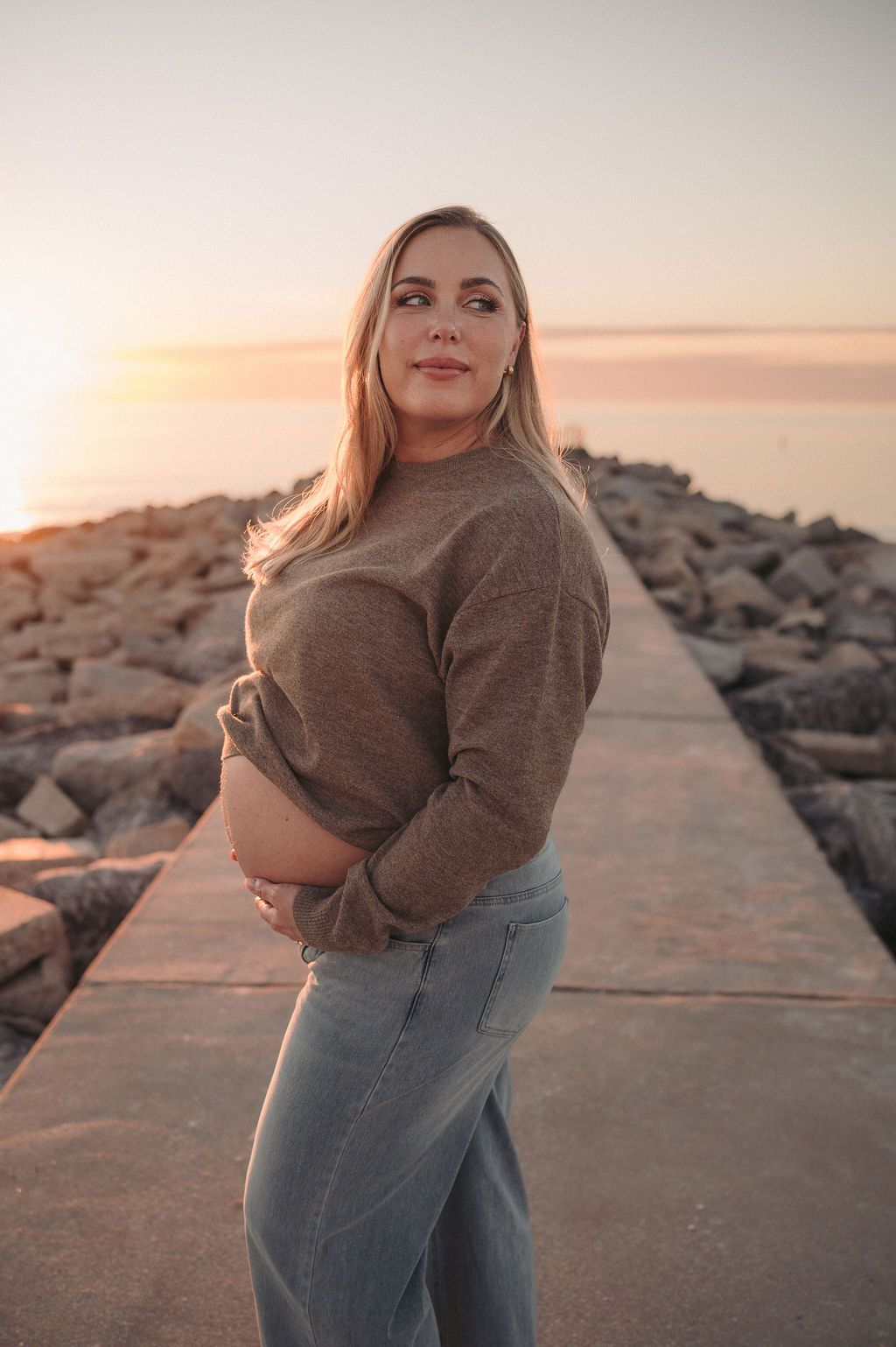 Pregnant mom standing on rocky beach lat sunset looking over her shoulder and showing her belly,