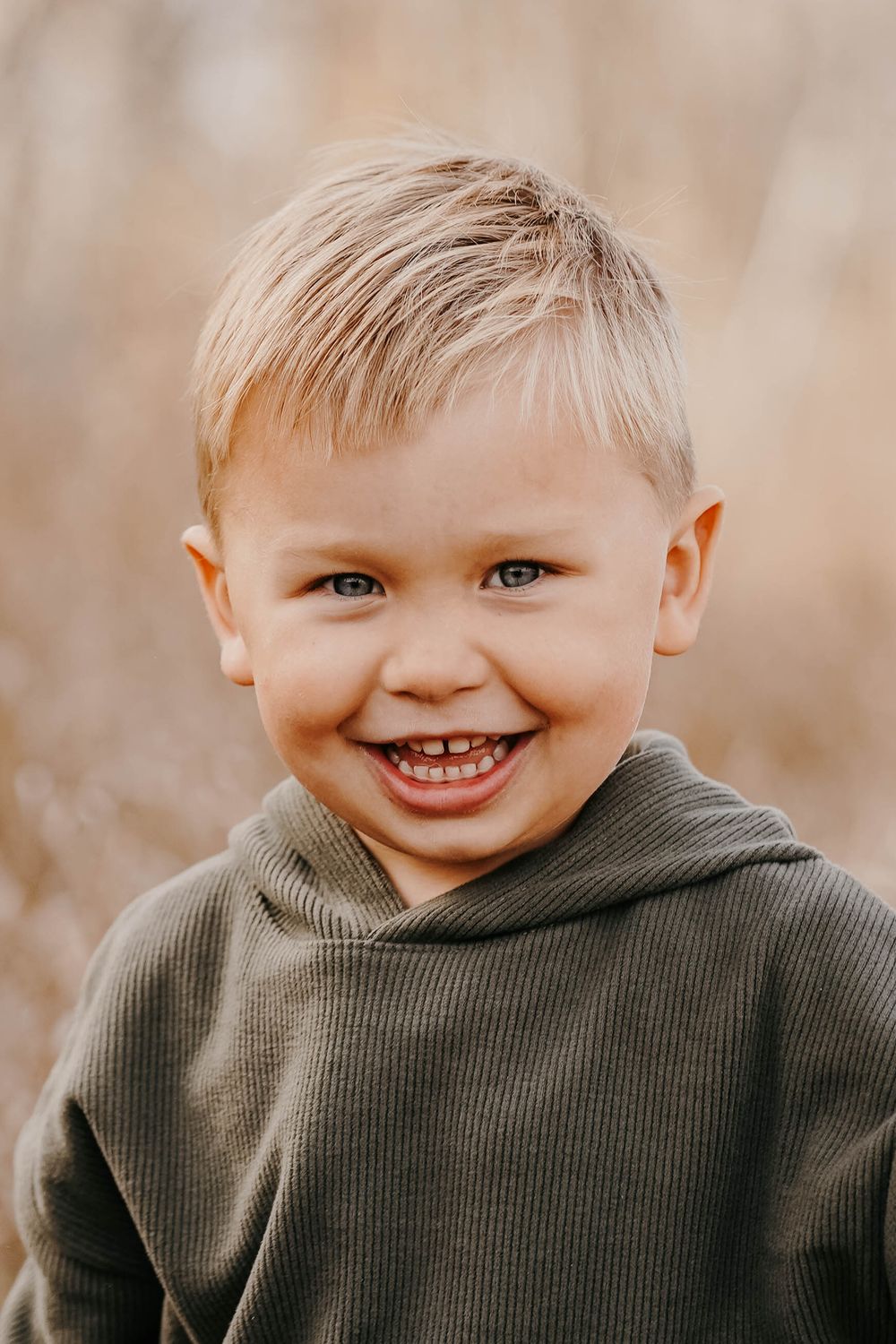A smiling young blonde boy with blue eyes wearing an olive green hoodie outdoors in a natural setting.