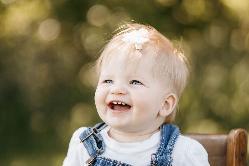 Smiling baby with a daisy in hair wearing denim overalls, sitting outdoors in warm natural light.
