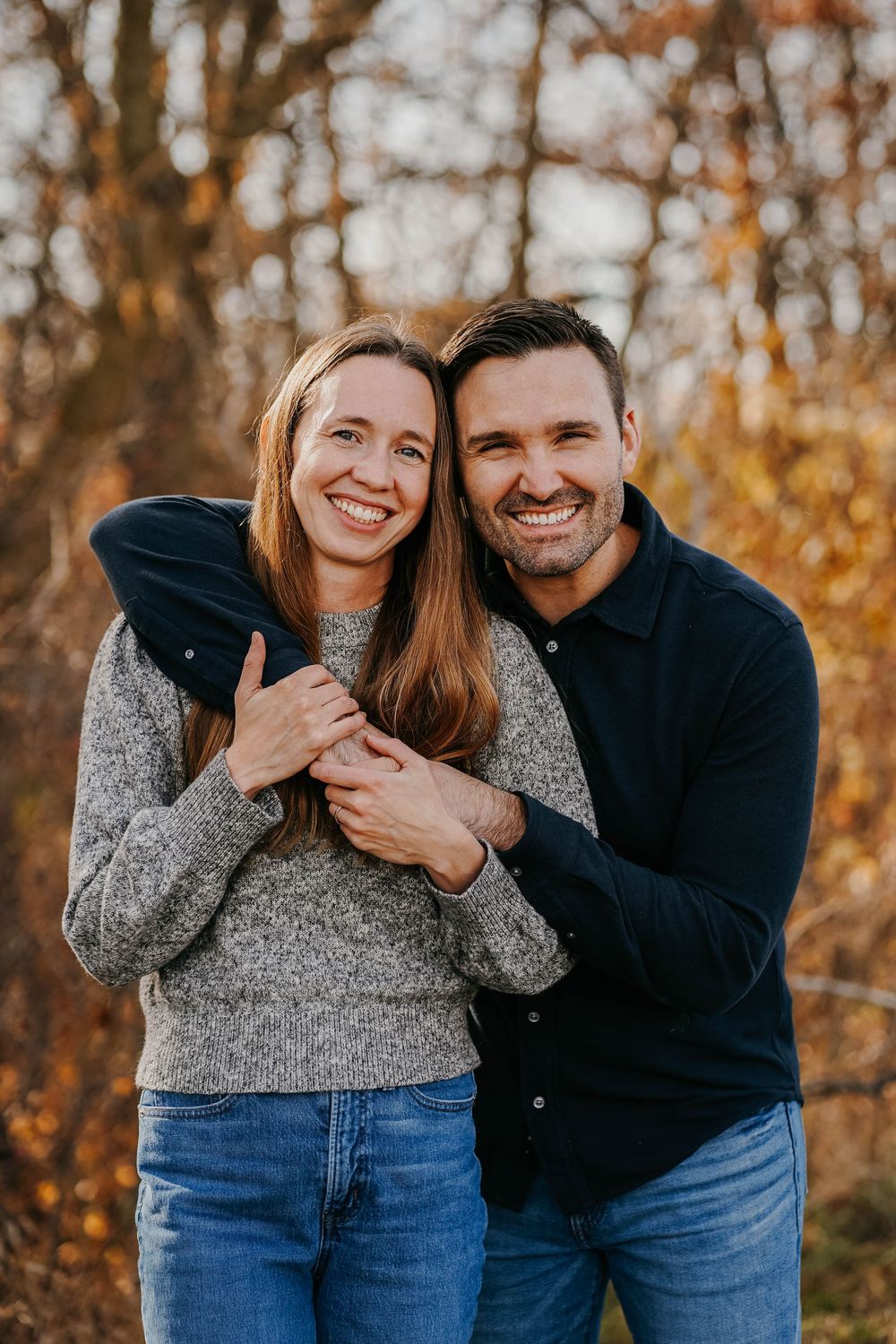 Smiling couple embracing outdoors in autumn, wearing casual jeans and sweaters with colorful fall foliage in background.