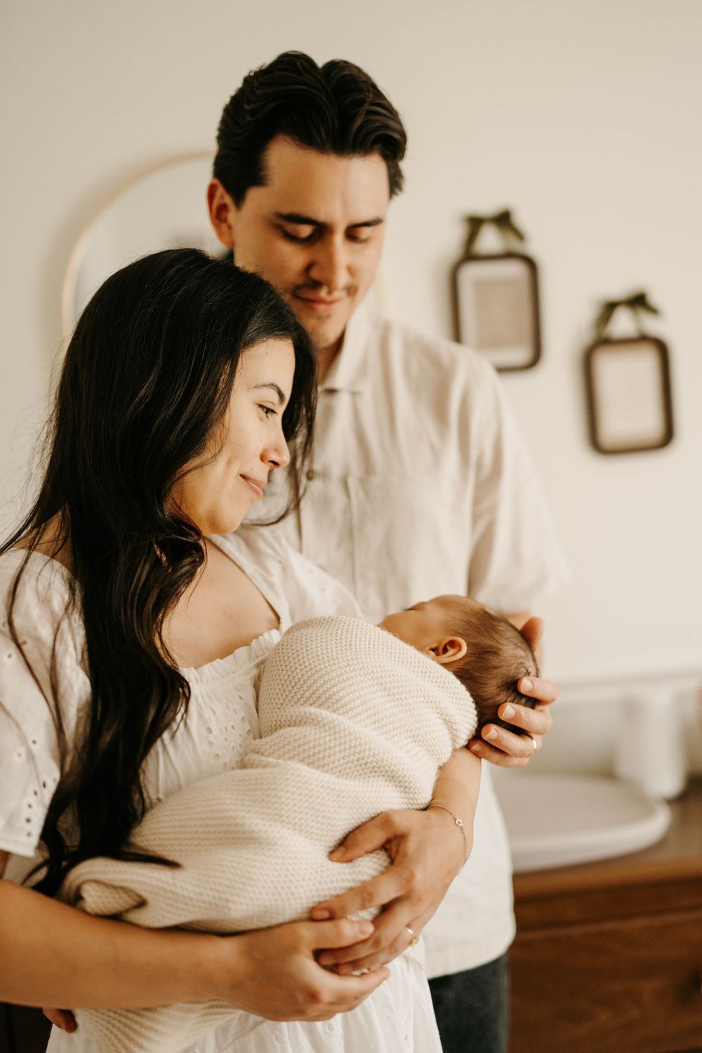 A couple in white clothing tenderly gazes at their newborn baby in a warmly lit nursery room.