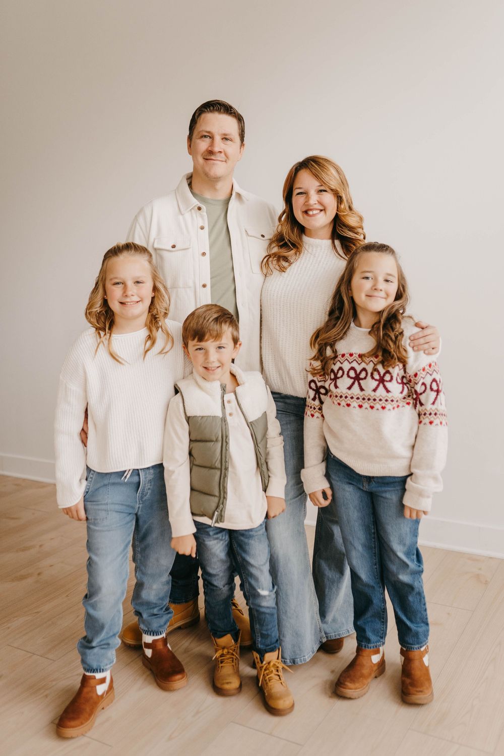 Family of five posing for a holiday portrait in coordinating neutral outfits and jeans on a light wood floor.